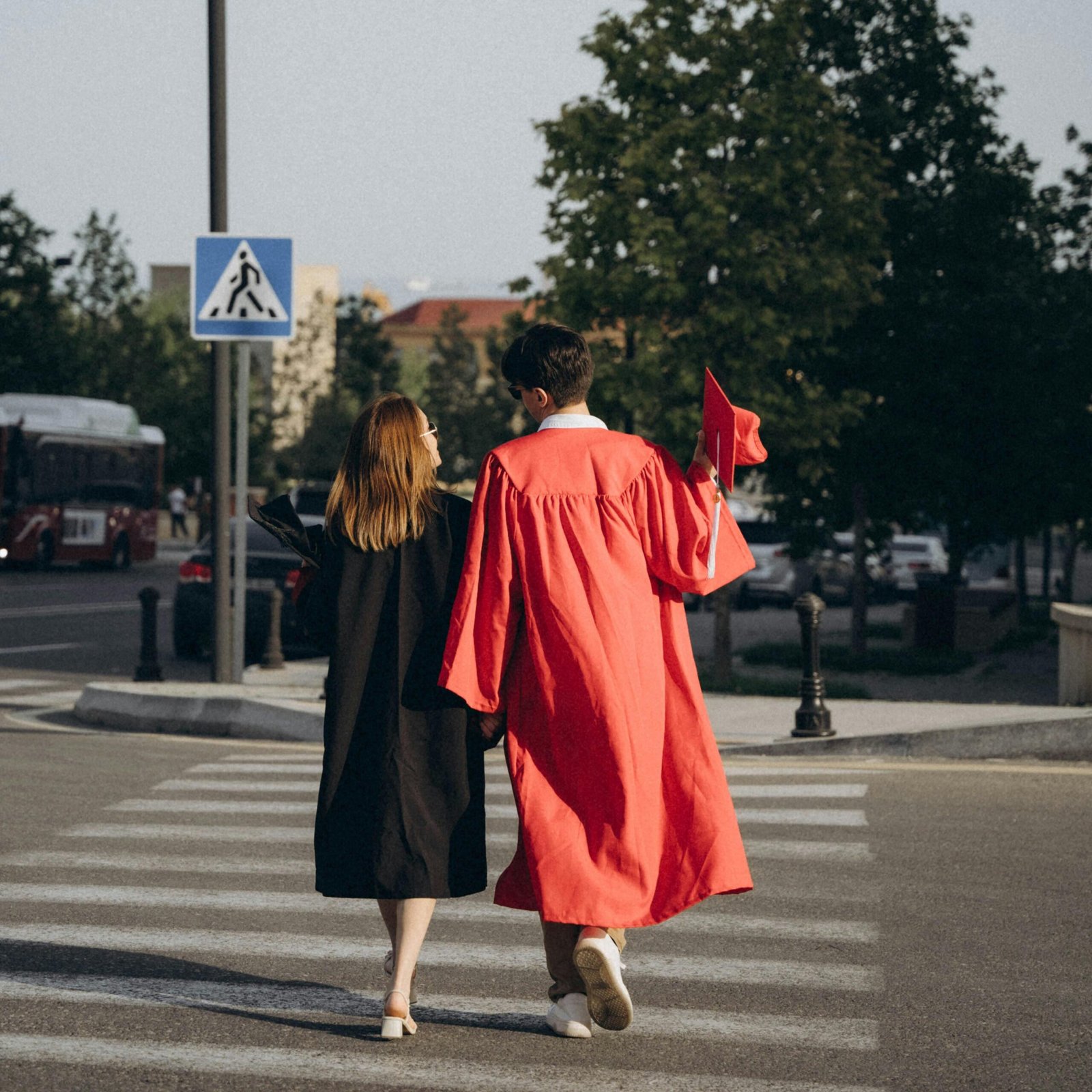 A pair of graduates wearing traditional gowns walk hand in hand across a city crosswalk celebrating their achievement.
